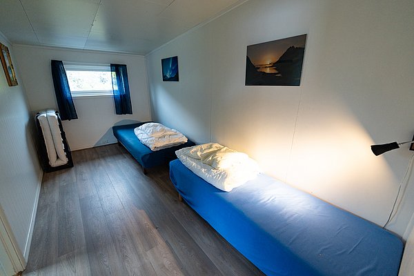 Gemütliches Schlafzimmer in Nordland, Norwegen, mit zwei Einzelbetten und minimalistischem Design. Helle Wände, blau bezogene Betten, Fenster mit blauen Vorhängen und dekorativen Landschaftsbildern an den Wänden. Ideal für ruhigen Angelurlaub.