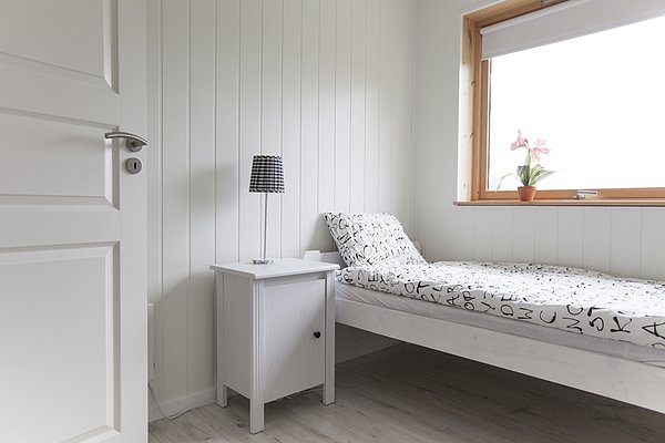 Ein helles, minimalistisches Schlafzimmer in Nord-Norwegen mit Einzelbett, Beistelltisch und Lampe. Blick aus dem Fenster auf eine blühende Pflanze. Ideal für einen ruhigen, erholsamen Angelurlaub.