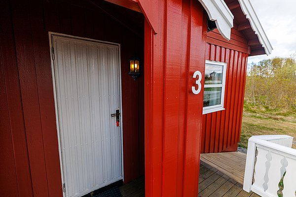 Einladendes rotes Ferienhaus mit weißer Tür und Nummer drei, typisch für die malerische Küstenregion Hitra, Norwegen. Perfekte Unterkunft für Angelurlauber auf der Suche nach Entspannung und Abenteuer in eindrucksvoller Natur.