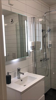 Modernes Badezimmer in Nord-Norwegen: weiße Fliesen, Glas-Duschkabine, Spiegelbeleuchtung, Waschbecken mit Armatur. Ideal für Erholung nach einem erlebnisreichen Angeltag. Perfekte Unterkunft für Angelreisen und Outdoor-Abenteuer in der skandinavischen Natur.