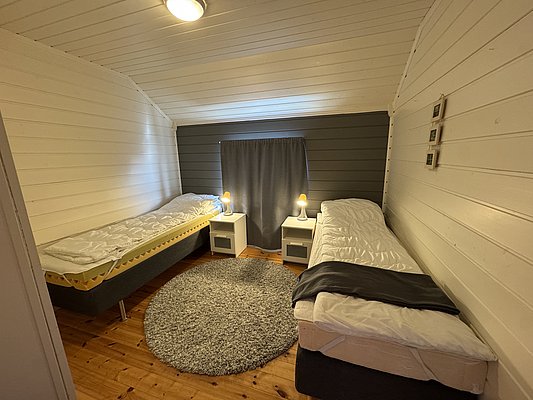 Gemütliches Schlafzimmer mit zwei Einzelbetten, Holzwänden und -boden, in Hitra, Norwegen. Ideal für Angelurlaub. Dezente Beleuchtung und neutraler Teppich schaffen eine einladende Atmosphäre für erholsame Nächte nach einem ereignisreichen Angeltag.