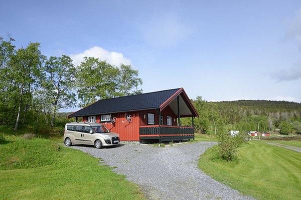 Rotes Holzhaus mit überdachter Terrasse, Auto davor geparkt. Umgeben von grüner Landschaft und Bäumen in Nordland, Norwegen. Ideal für Angelurlaub und Outdoor-Abenteuer. Ruhige, idyllische Umgebung perfekt zum Entspannen und Naturerleben.