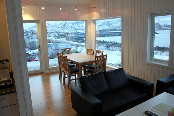 Helles Wohnzimmer in Nord-Norwegen mit großer Fensterfront und Ausblick auf verschneite Berge und das Meer. Gemütliche Einrichtung mit Esstisch und Sofa, ideal für Angelurlaub geeignet. Perfekte Kulisse für Entspannung und Naturerlebnis.