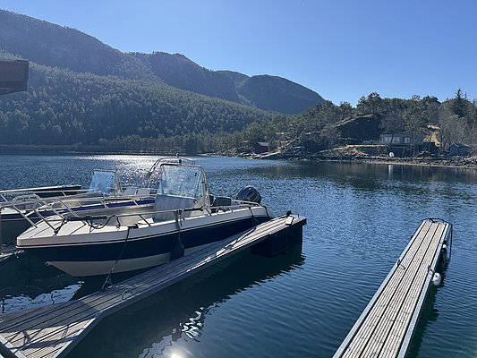 Angelboote liegen an einem ruhigen Steg, umgeben von atemberaubender Landschaft in Hitra, Norwegen. Die klare Wasseroberfläche spiegelt die üppigen Wälder und Berge wider, perfekt für einen Angelurlaub in unberührter Natur.