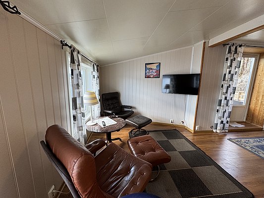 Gemütlicher Wohnbereich in einem Ferienhaus in Hitra, Norwegen. Ledersessel, Flachbild-TV und Fenster mit Vorhängen bieten Komfort und Entspannung nach einem langen Angeltag. Ideal für Angelreisende, die Erholung suchen.