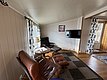 Gemütlicher Wohnbereich in einem Ferienhaus in Hitra, Norwegen. Ledersessel, Flachbild-TV und Fenster mit Vorhängen bieten Komfort und Entspannung nach einem langen Angeltag. Ideal für Angelreisende, die Erholung suchen.