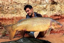Ein Mann hält stolz einen großen Karpfen vor einer lehmigen Wand in die Kamera.