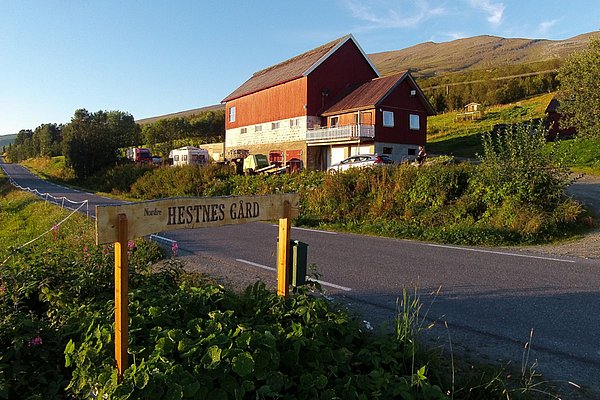 Hof in Nordnorwegen bei Sonnenuntergang, umgeben von grüner Natur und Bergen, mit rustikalem Gebäude und Hinweisschild „Nordre Hestnes Gård“ an einer ruhigen Landstraße. Ideal für idyllische Angelreisen und Naturerlebnisse.