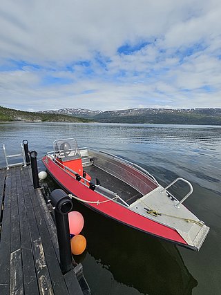 Ein rotes Boot ist an einem Holzsteg festgemacht, umgeben von bunten Bojen, mit einem ruhigen See und bewölktem Himmel im Hintergrund.